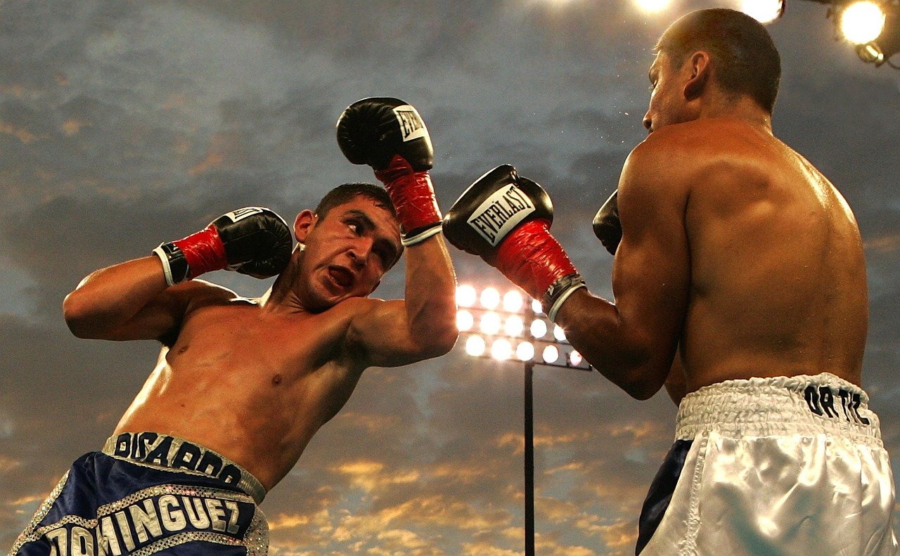 Two shirtless boxers in action in a well-lit outdoor ring during sunset, wearing red gloves and focused expressions.