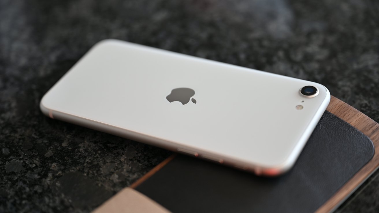White smartphone with a silver logo lies on a dark, speckled surface, its camera lens facing upward, next to a wooden object.