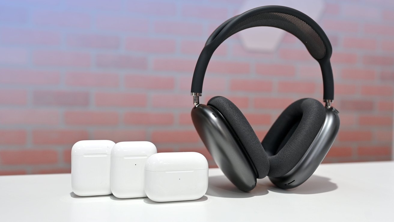 Three white wireless earbuds cases next to large black over-ear headphones on a white surface, with a brick wall background.