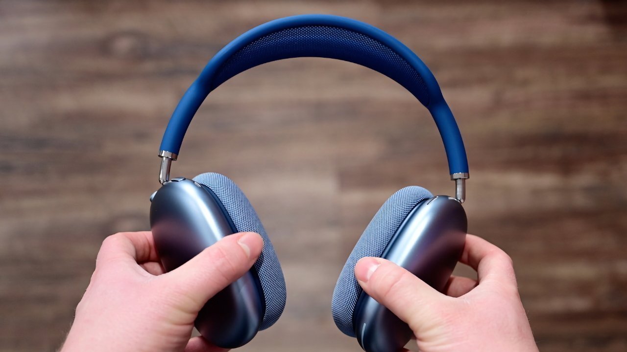 Hands holding blue over-ear headphones with mesh headband, against a wooden floor background.